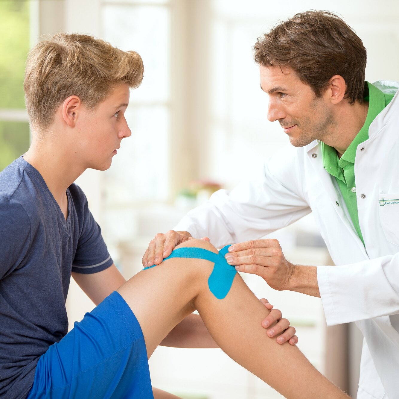 Junger Arzt behandelt einen Jungen am Knie mit Kinesio-Tape.