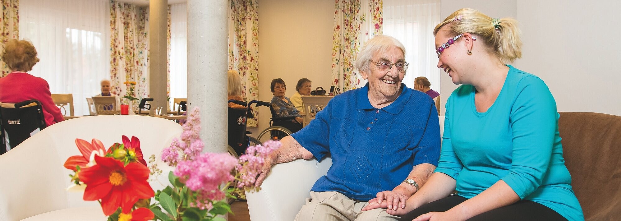 Gemeindepflegehaus Brieselang: Wir ermöglichen gesellschaftliche Teilhabe sowie ein selbstbestimmtes und zufriedenes Leben. Eine junge und eine alte Frau sitzen gemeinsam in einem Gemeinschaftsraum auf einer Couch und lachen sich an. Im Hintergrund sind weitere Bewohnerinnen und Bewohner zu sehen. Auf einem Tisch im Vordergrund des Bildes steht eine Vase mit einem bunten Blumenstrauß.
