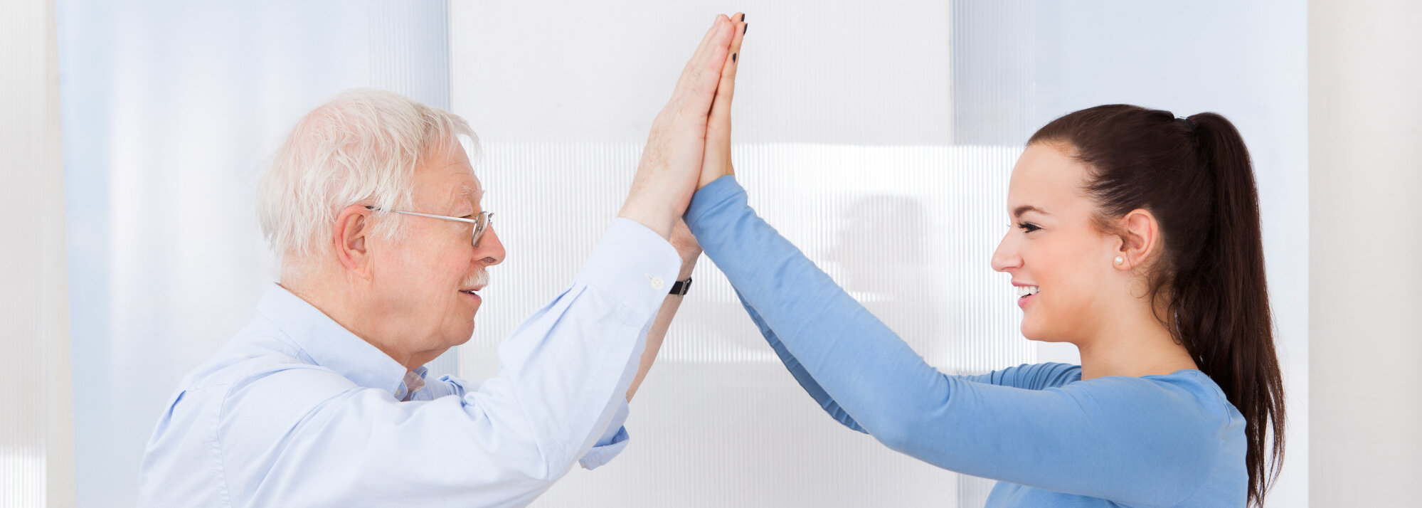 Seitliche Aufnahme eines älteren Herren in weißem Hemd und einer jungen Frau in einem hellblauem Langarm-Shirt. Beide lächeln sich an, strecken ihre Hände in die Höhe und berühren sich gegenseitig mit den Handflächen.