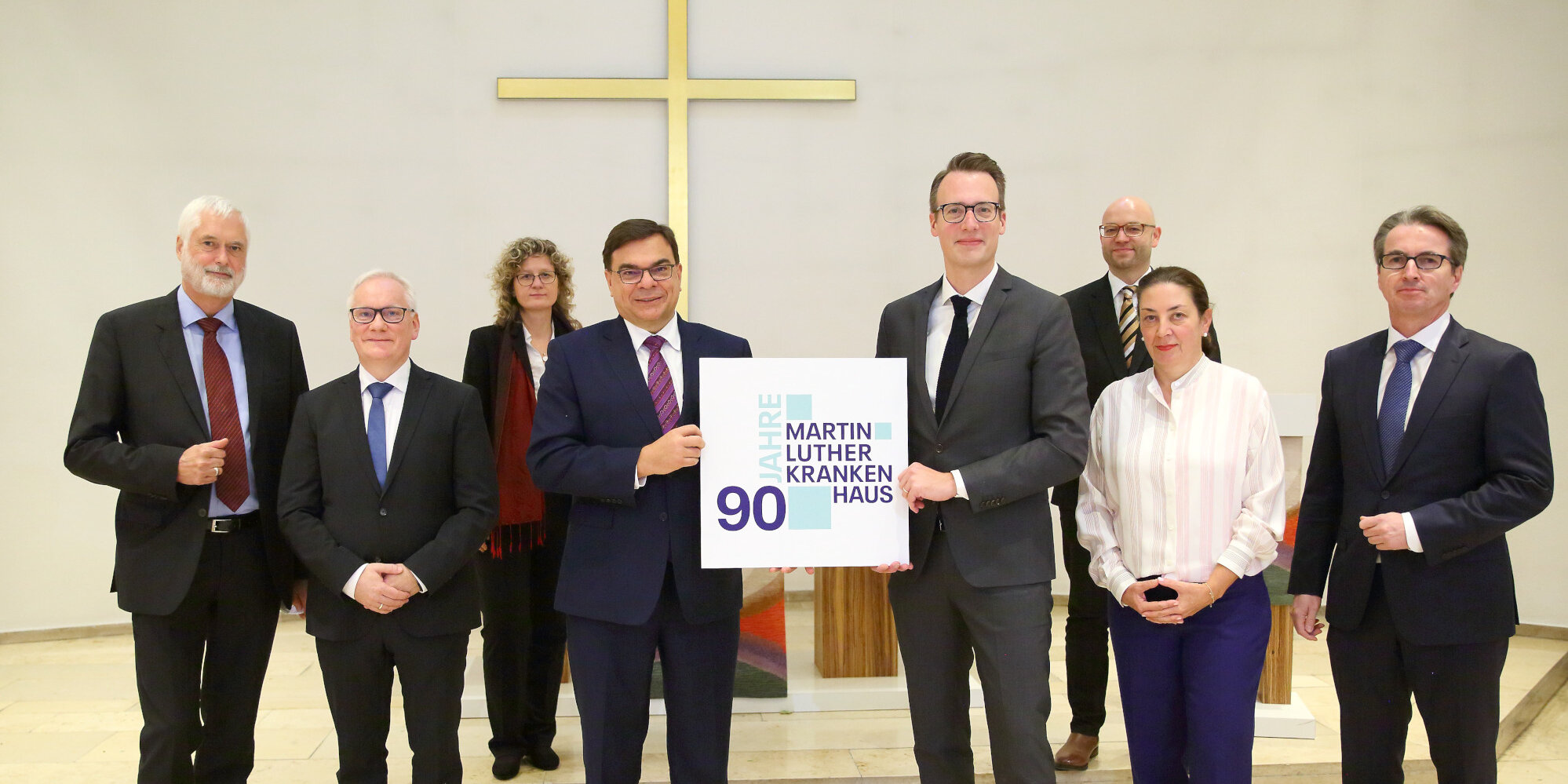 Acht Männer und Frauen stehen in der Kapelle und halten ein Schild mit der Aufschrift "90 Jahre Martin Luther Krankenhaus".