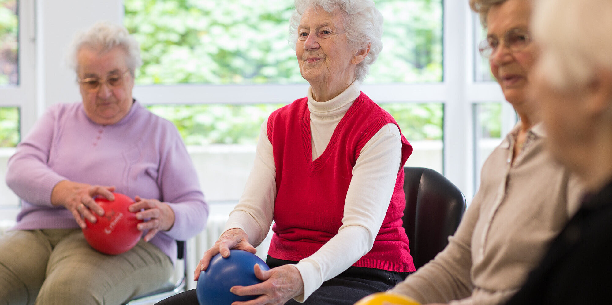 Vier Rentnerinnen bei einem geselligen Ballspiel, sitzend auf Stühlen. In den Händen halten sie jeweils einen kleinen, bunten Kunststoffball.