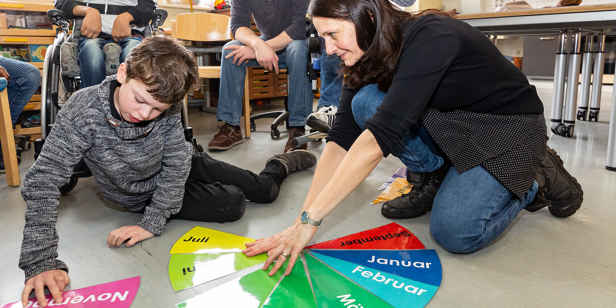 Wohlfühlatmosphäre: Kleine Klassen und eine angenehme Lernumgebung Ein Schüler und eine Lehrerin sitzen auf dem Fußboden; im Hintergrund weitere Schüler sowie ein Betreuer. Sie lernen anhand bunter Lernkarten die Monate eines Jahres.