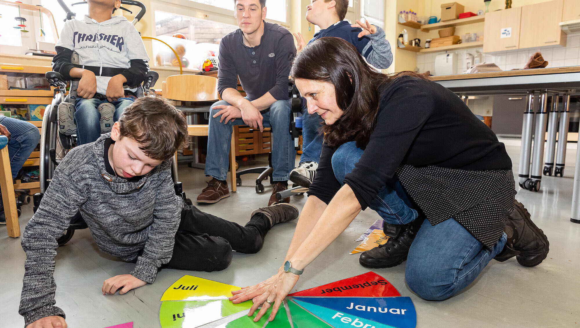 Wohlfühlatmosphäre: Kleine Klassen und eine angenehme Lernumgebung Ein Schüler und eine Lehrerin sitzen auf dem Fußboden; im Hintergrund weitere Schüler sowie ein Betreuer. Sie lernen anhand bunter Lernkarten die Monate eines Jahres.