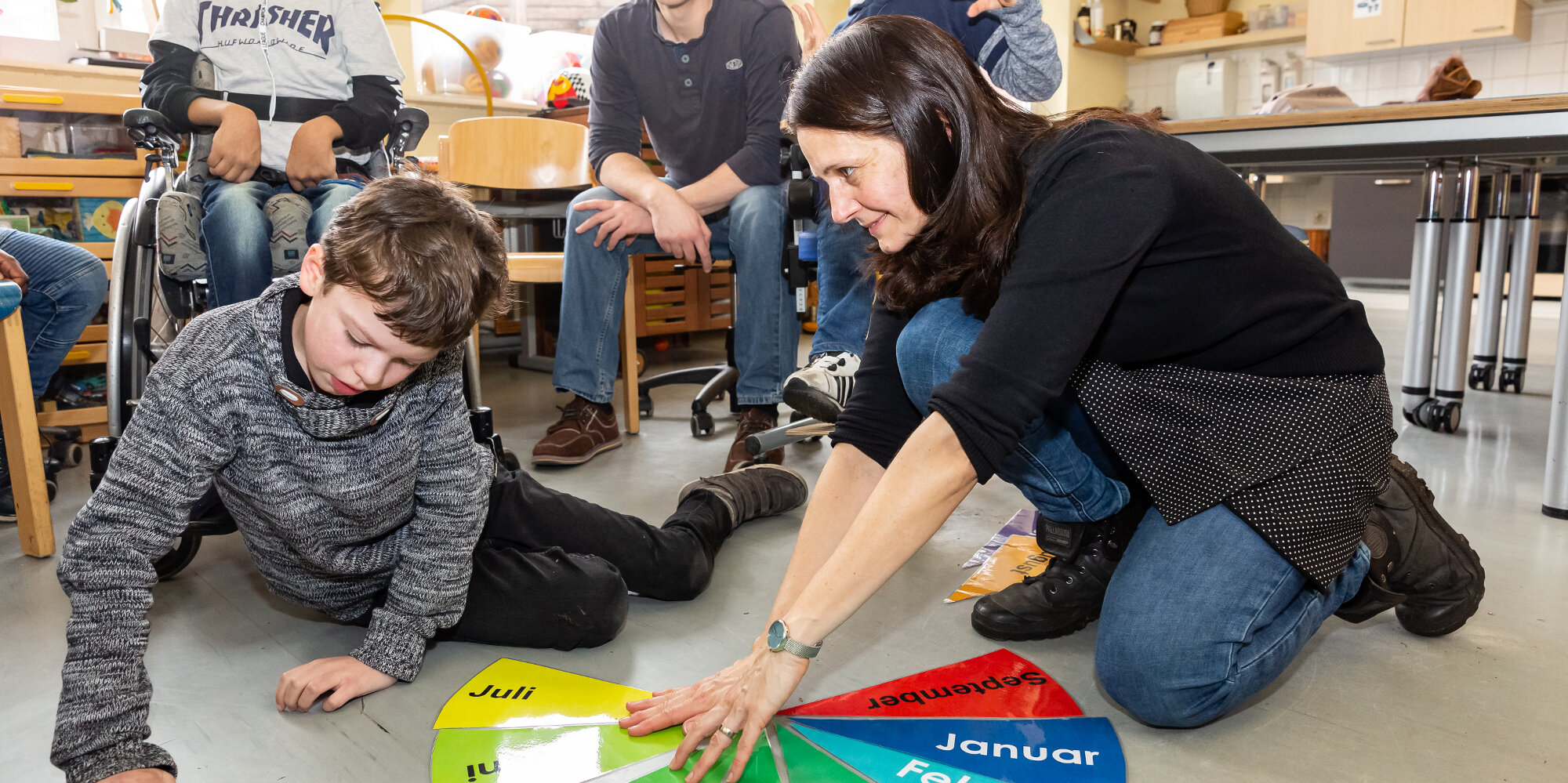 Ein Schüler und eine Lehrerin sitzen auf dem Fußboden; im Hintergrund weitere Schüler sowie ein Betreuer. Sie lernen anhand bunter Lernkarten die Monate eines Jahres.