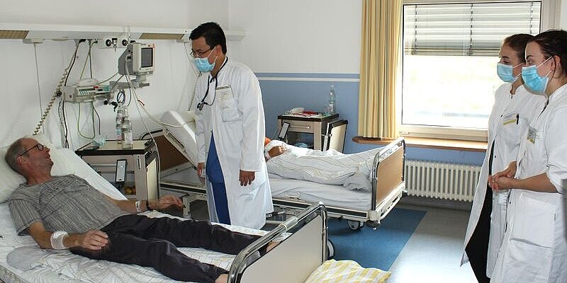 Ein Arzt spricht mit einem Patienten am Patientenbett. Zwei Teilnehmende der Summer School schauen zu.