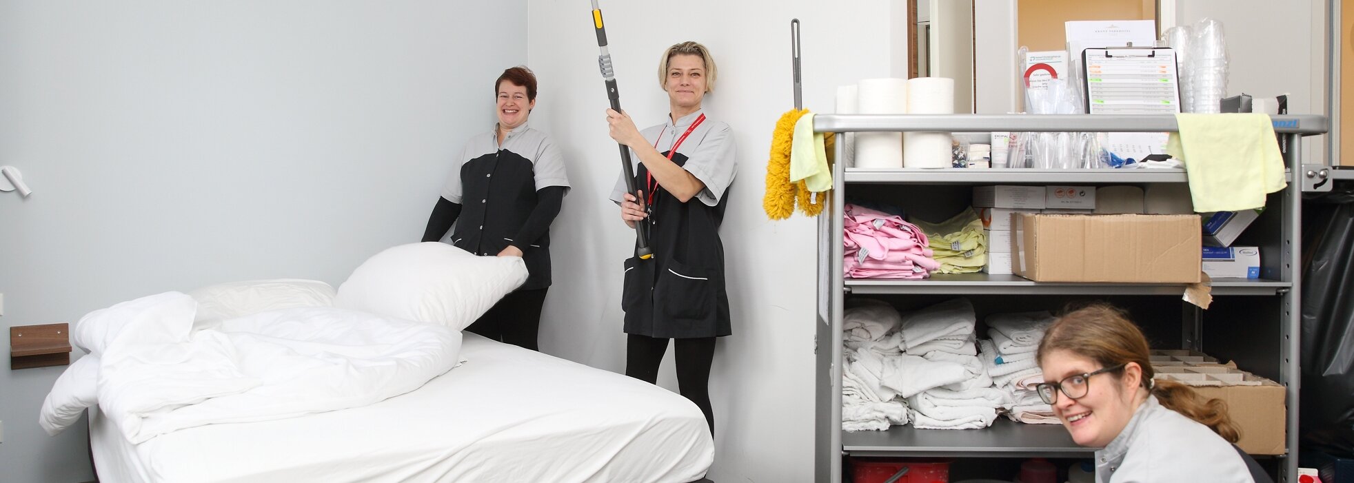 Drei gut gelaunte Mitarbeiterinnen posieren während der Reinigungsarbeiten in einem Hotelzimmer. Rechts im Bild steht ein großer Rollwagen mit Utensilien.