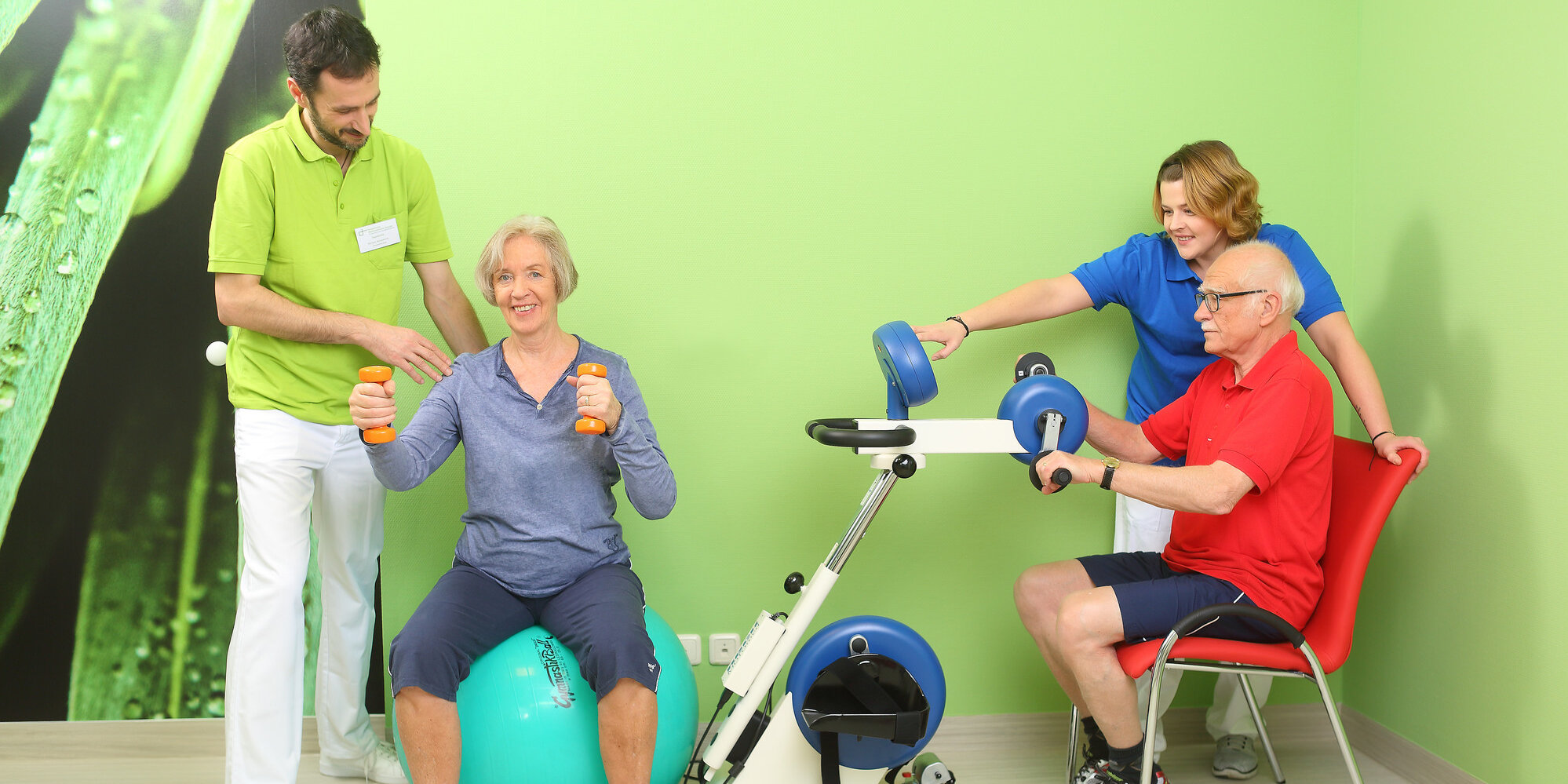 Zwei ältere Menschen trainieren mit Trainingsgeräten und werden von zwei Mitarbeitenden der Physiotherapie unterstützt.