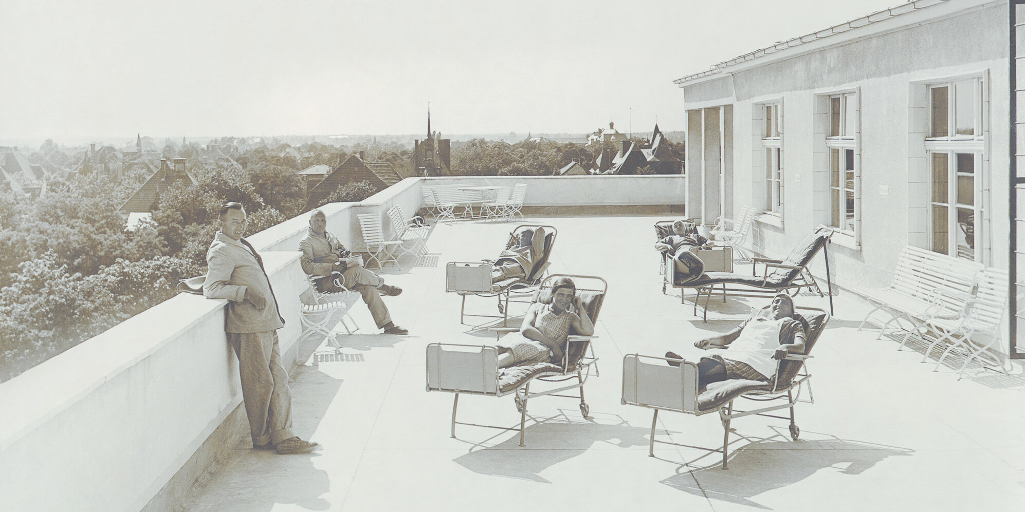 Vier Patientinnen im Liegestuhl auf der Dachterrasse. Ein Mann steht am Rand des Daches und ein Mann sitzt auf einem Stuhl.