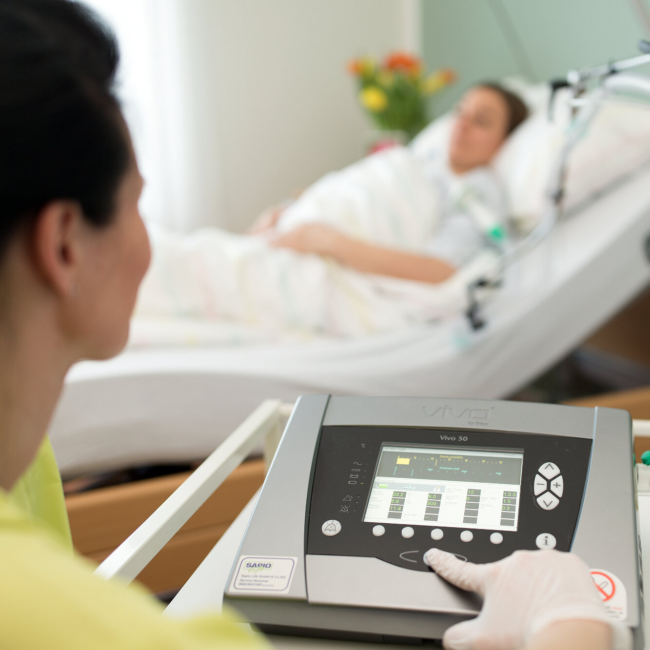Eine Pflegerin steuert ein medizinisches Atemgerät. Im Hintergrund ist eine Patientin im Pflegebett zu sehen.