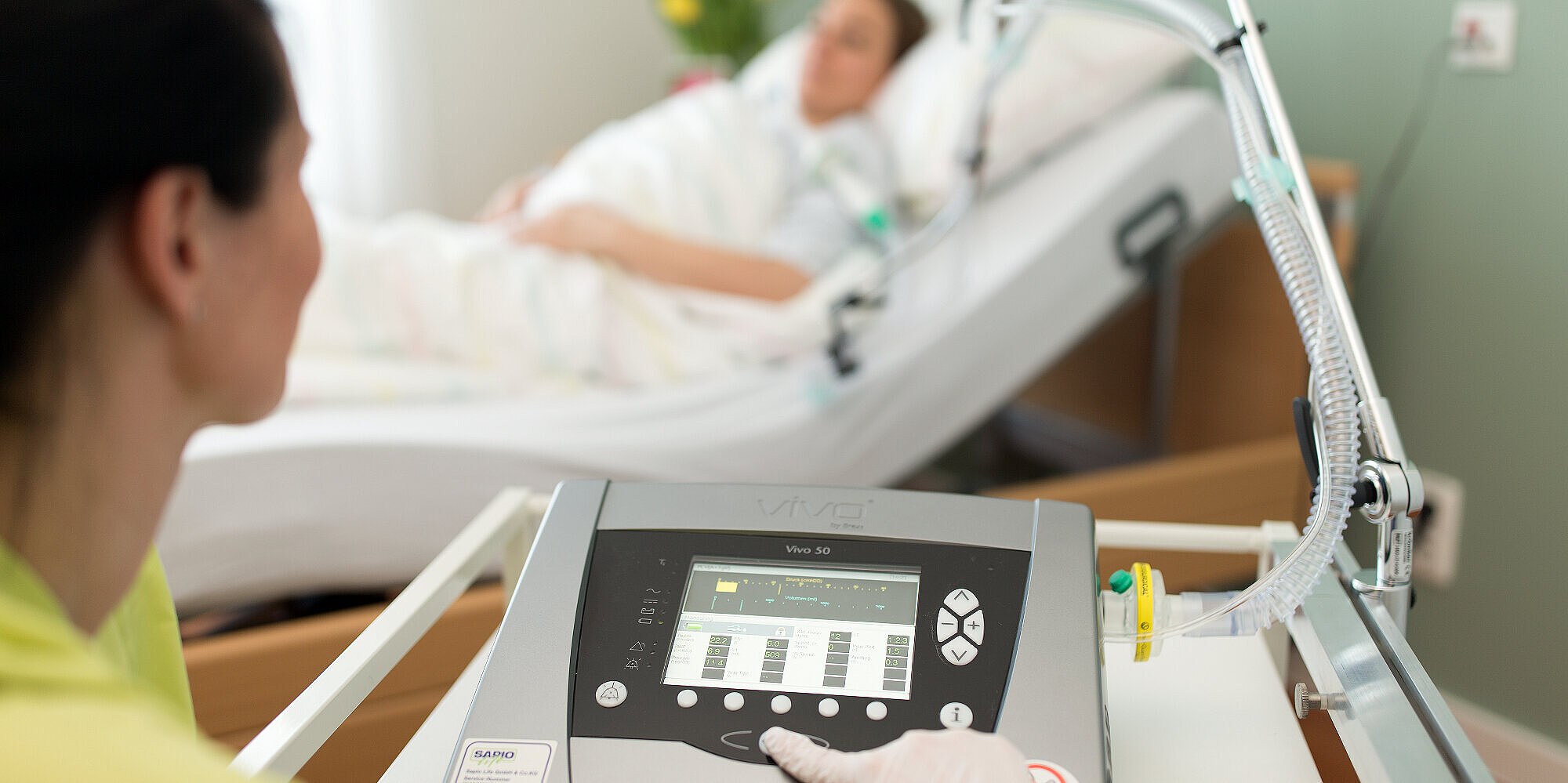 Eine Pflegerin steuert ein medizinisches Atemgerät. Im Hintergrund ist eine Patientin im Pflegebett zu sehen.