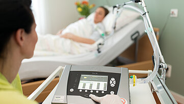 Eine Pflegerin steuert ein medizinisches Atemgerät. Im Hintergrund ist eine Patientin im Pflegebett zu sehen.