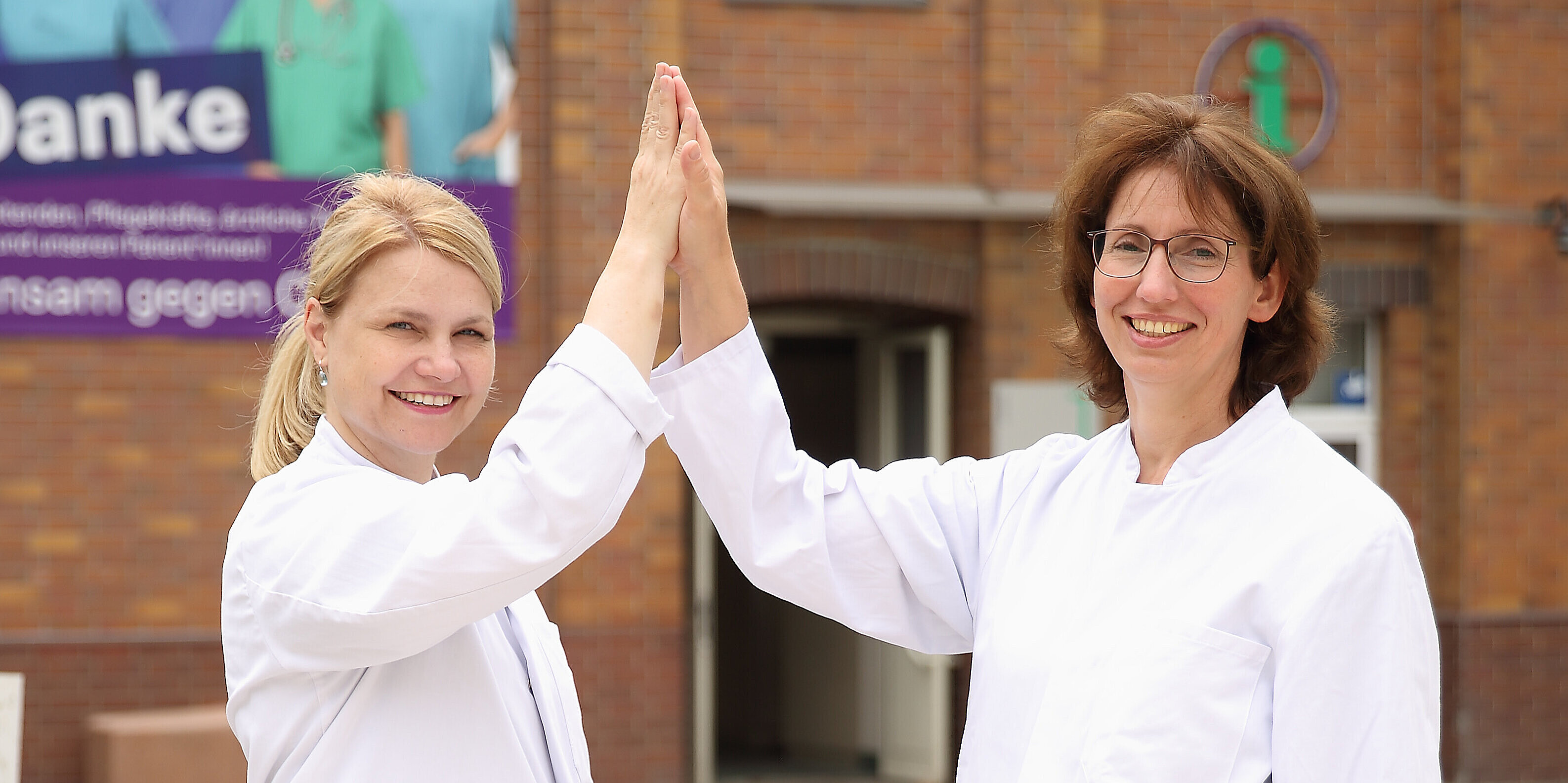 Zwei Chefärztinnen im Jobsharing-Modell geben sich ein High-Five vor der Klinik – erfolgreiche geteilte Führung als modernes Arbeitszeitmodell für Ärzte*Ärztinnen im Krankenhaus.