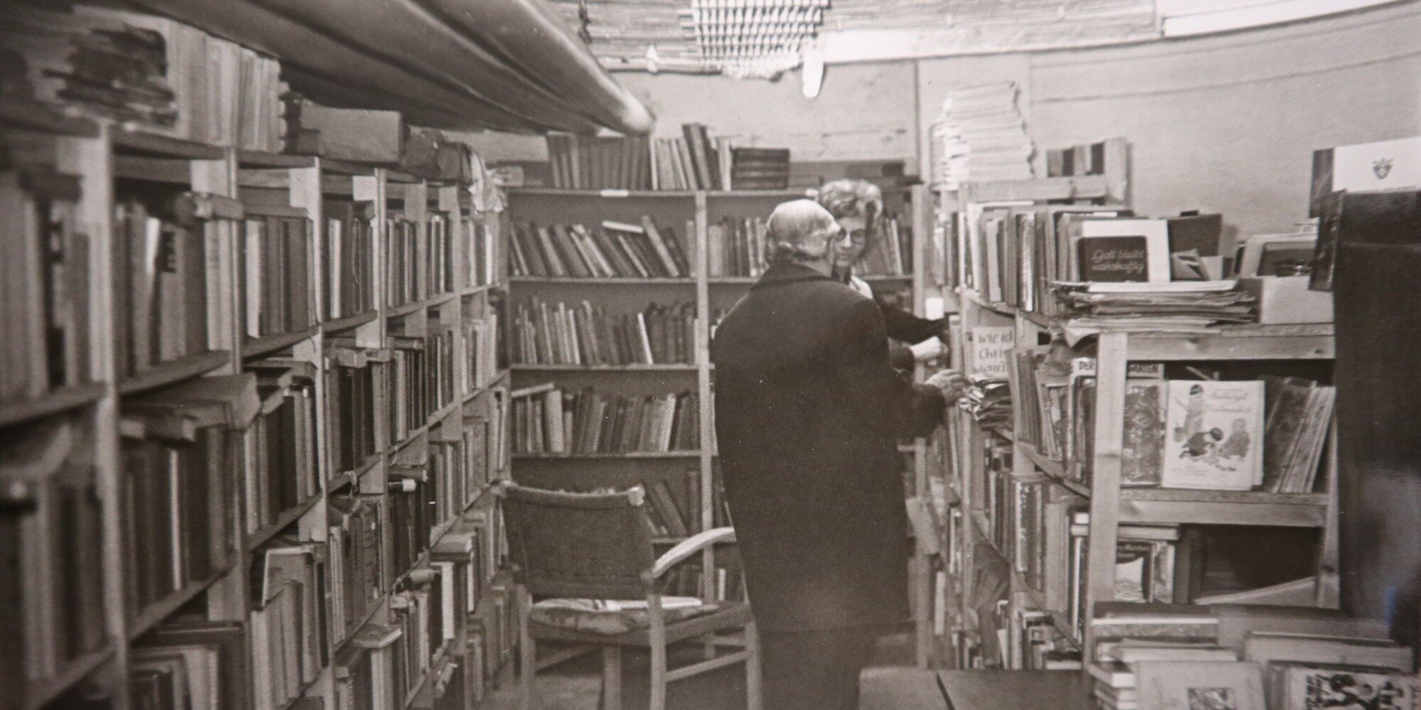Schwarz-Weiß-Aufnahme eines Raumes im ehemaligen Spendenkeller mit vielen Regalwänden, in denen Bücher stehen. Eine Frau und ein Mann schauen gemeinsam nach Büchern.