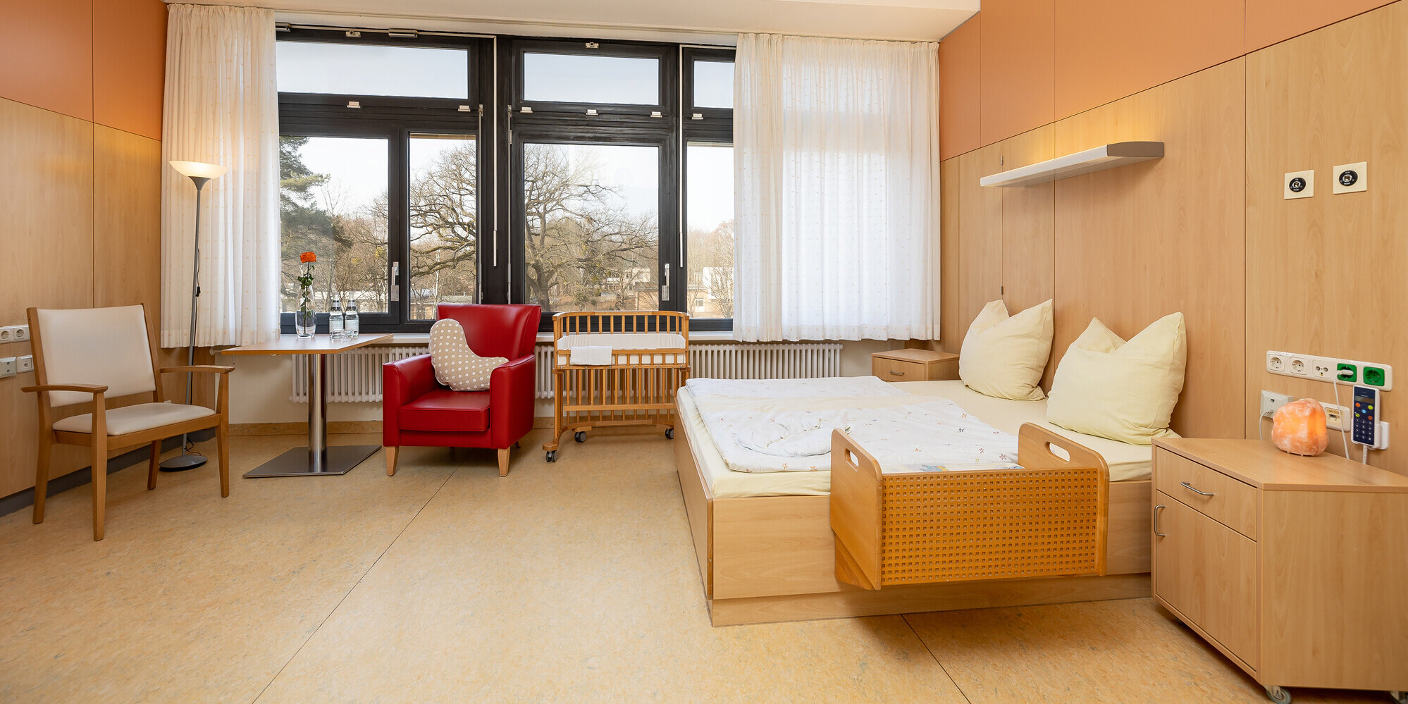 Großer heller Raum mit großen Fenstern, breitem Bett, Sitzmöglichkeiten und Babybett.