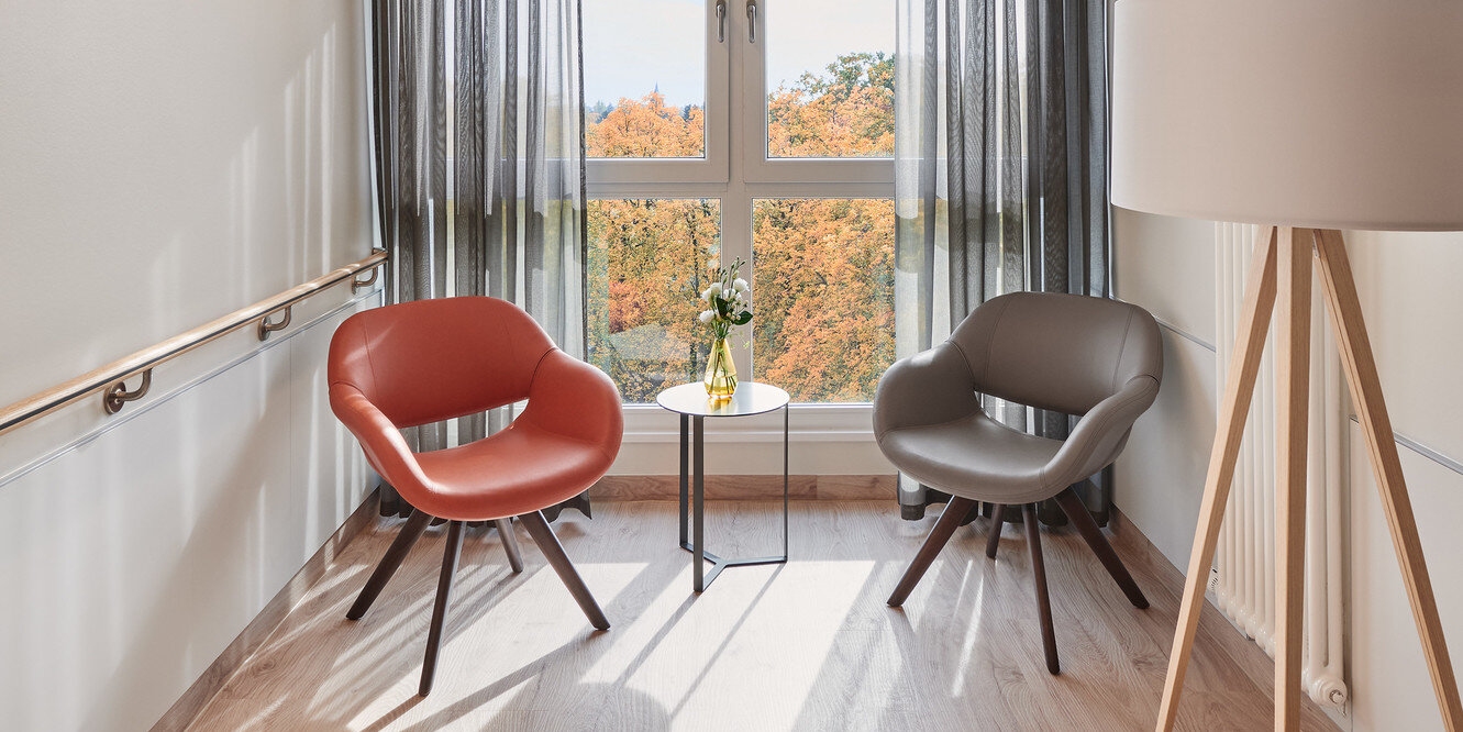 Blick in einen hellen Raum mit großem Fenster, einer kleinen Sitzecke inklusive moderner Beleuchtung.