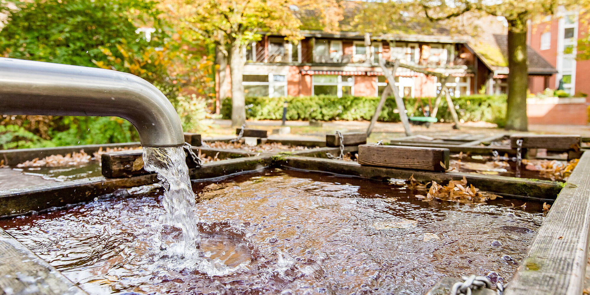 Aus einem Wasserhahn fließt Wasser in ein Holzbecken. Gebäude und eine Schaukel im Hintergrund.